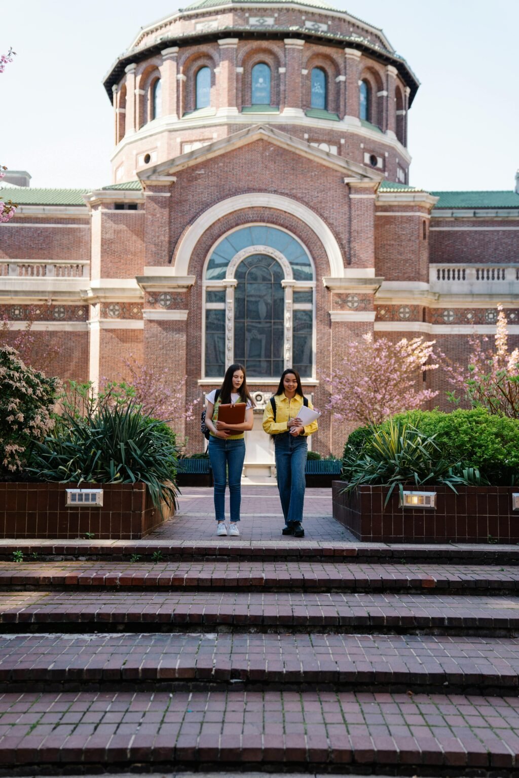 Two young women walking past a historic university building on a sunny day.